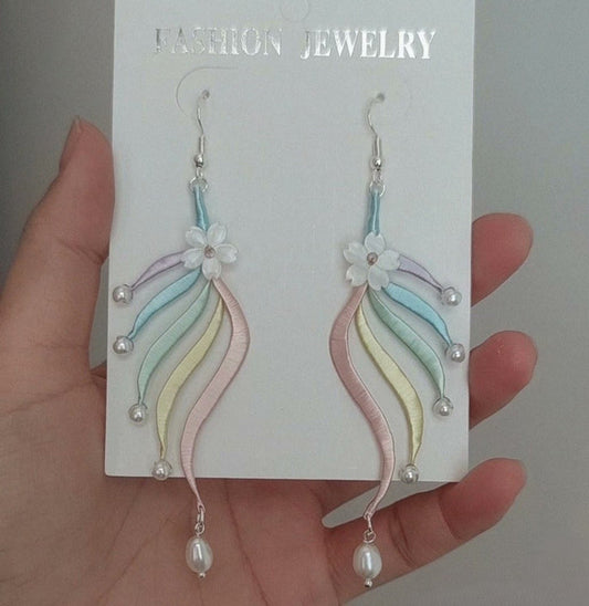 A pair of handmade Chanhua earrings featuring pastel silk tassels and a white flower, held against a white background.