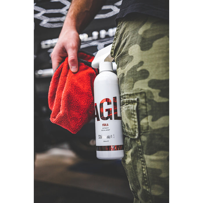 Close-up of a person holding a red microfiber towel and a bottle of Stjärnagloss Pärla spray sealant, with camo-patterned pants visible in the foreground.