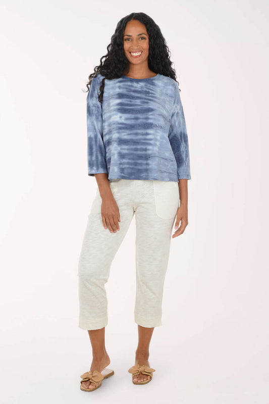 A woman modeling a blue and navy tie-dye long-sleeve top with off-white cropped pants and tan slide sandals, standing against a plain white studio background.